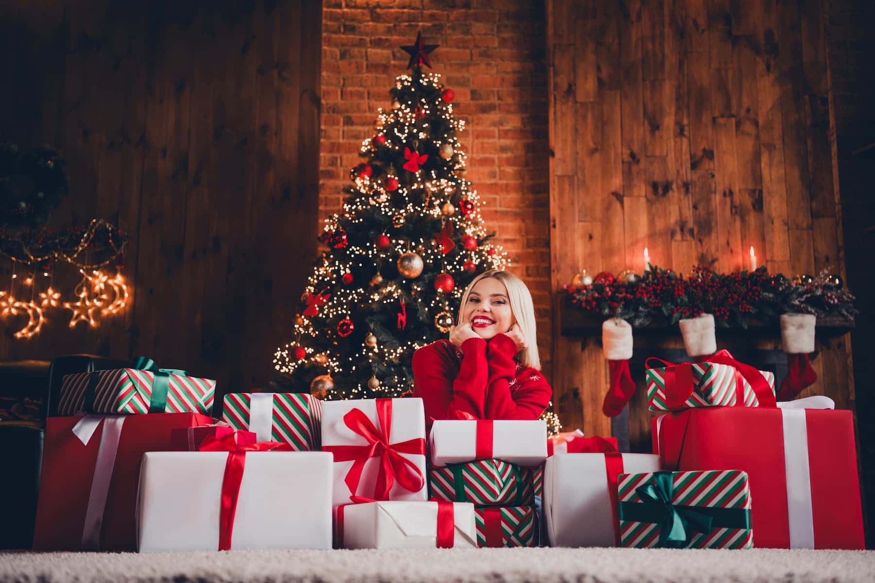 Full body photo of a young woman in a red sweater surrounded by a pile of gifts under the Christmas tree, showcasing Christmas gift ideas for friends and family.