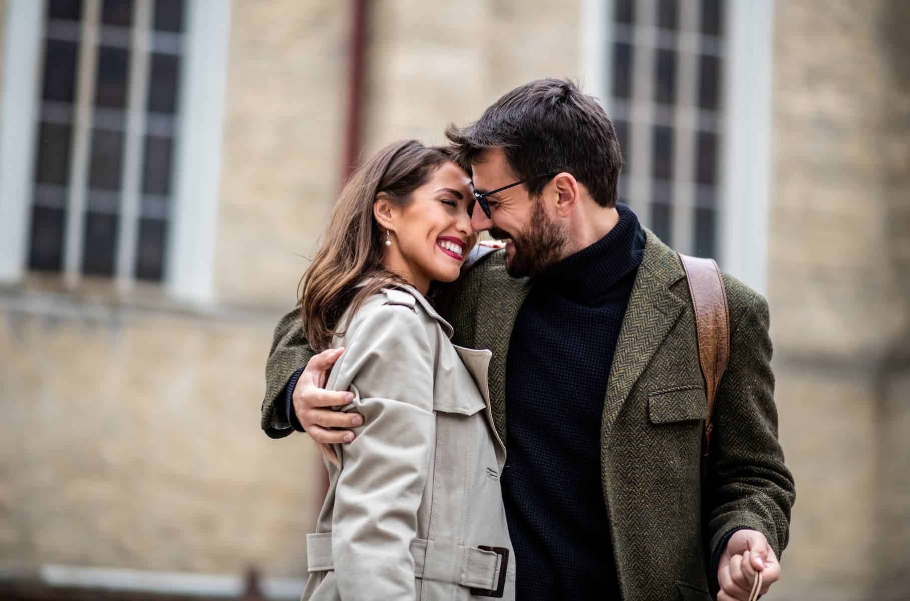 A couple embrace and smile in front of a building, symbolizing connection and closeness like quantum entanglement.