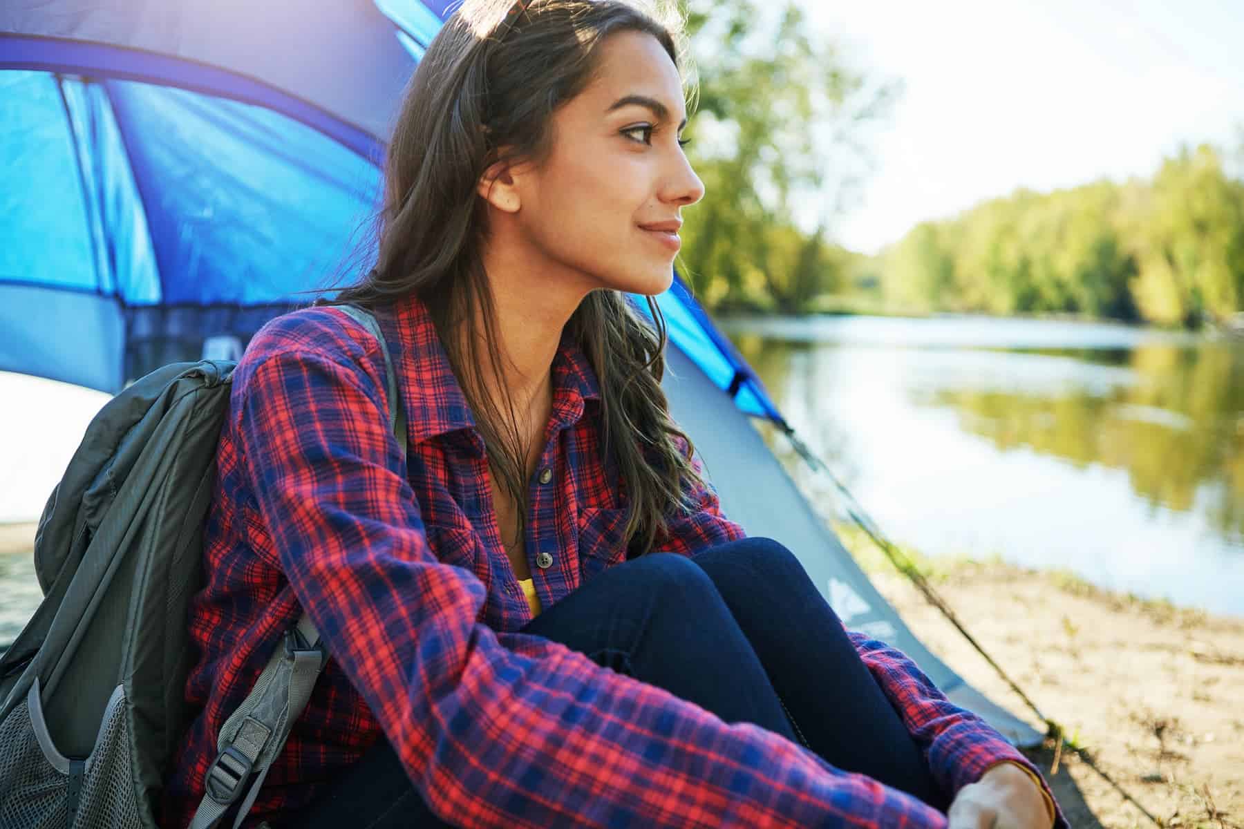 A woman wearing a plaid shirt sits in front of a tent with a backpack, looking out over a calm lake surrounded by trees.