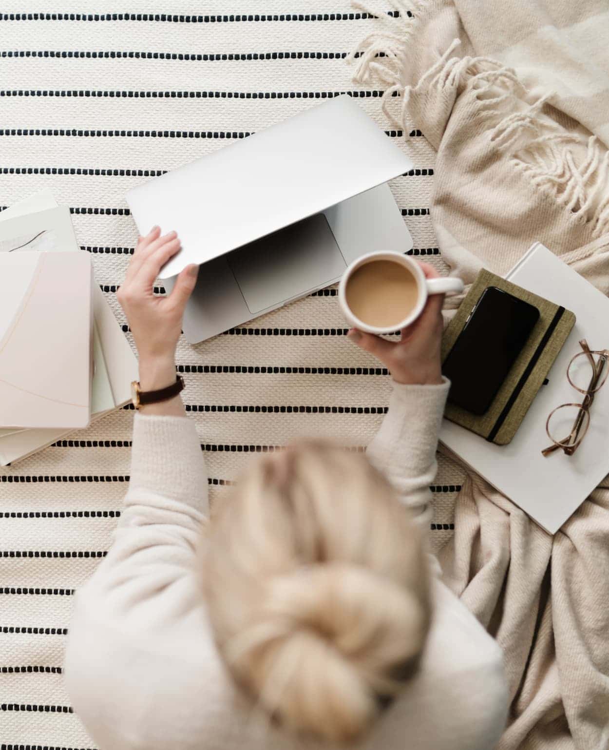 A woman opens her laptop while holding a warm cup of coffee, surrounded by neutral tones and soft fabric textures that create a quiet, mindful sense of focus and ease.