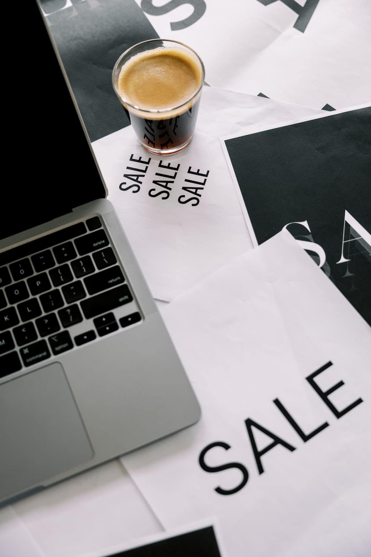 A laptop and a glass of coffee sit on papers with the word "SALE" printed on it, highlighting Black Friday deals.