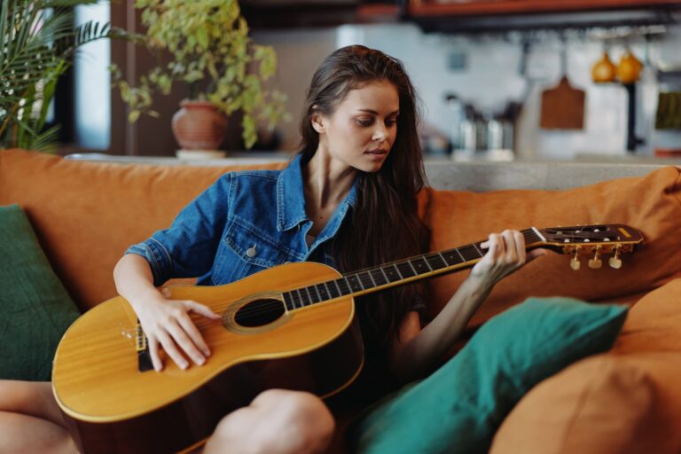 A woman practices playing the guitar while sitting on a couch at home.