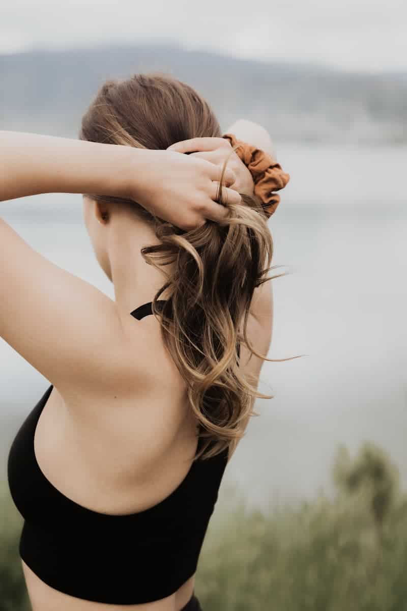 A woman with brown hair ties her hair up with a rust-colored scrunchie, set against a blurred backdrop of water and hills, reflecting a focused and intentional mindset.