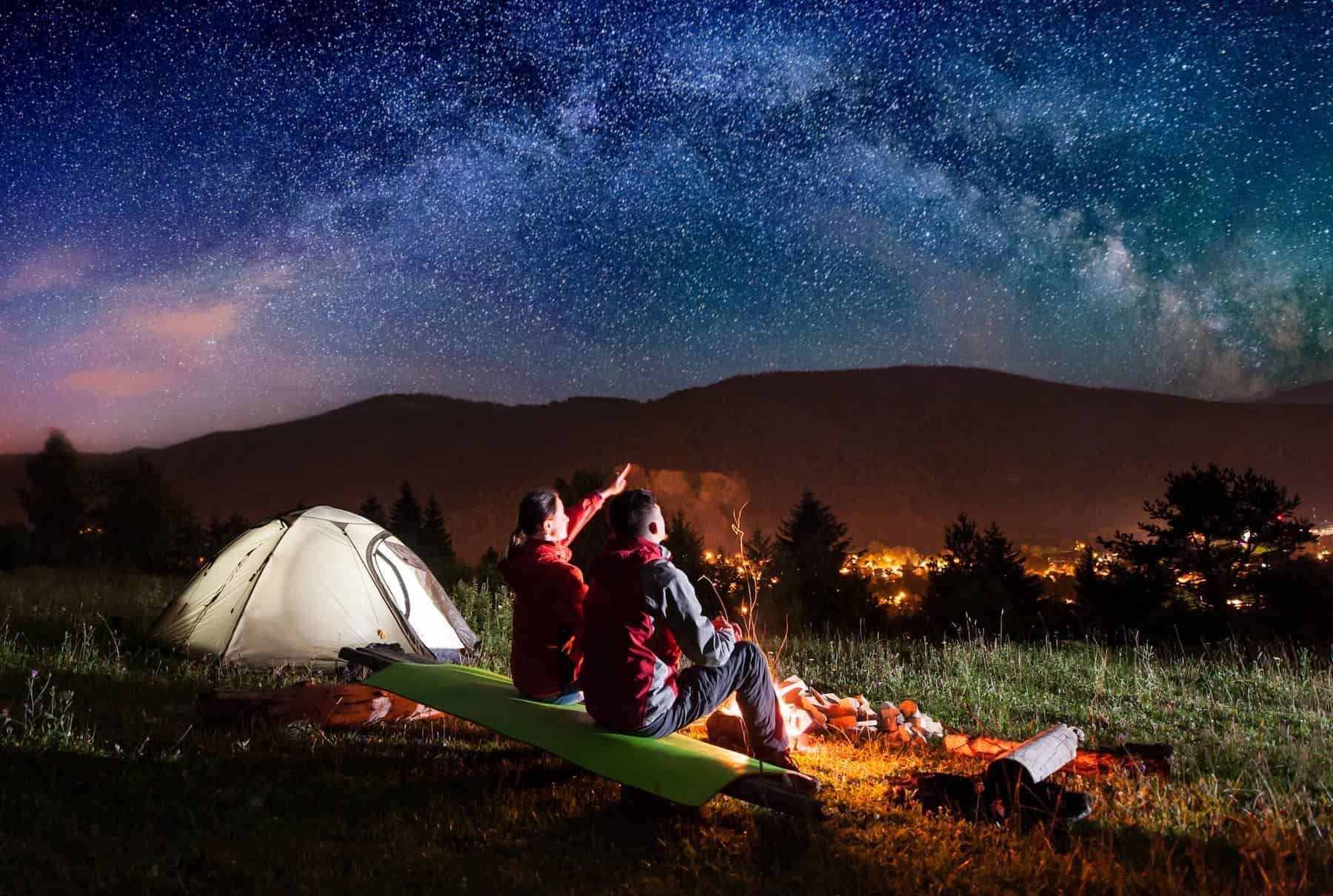 Two people sitting by a campfire under a starry night sky with tent and town lights, contemplating the mysteries of quantum physics.
