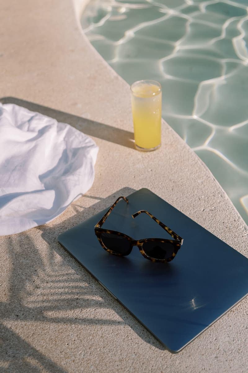 A pair of tortoiseshell sunglasses rests on a dark blue surface beside a glass of yellow liquid and a white towel by a swimming pool, evoking a quiet moment of self-reflection and stillness.