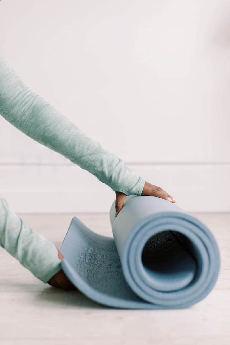 A woman in a light blue long-sleeved shirt rolls up a light blue yoga mat on a light-colored floor, embracing a quiet moment of wellbeing and self-care.