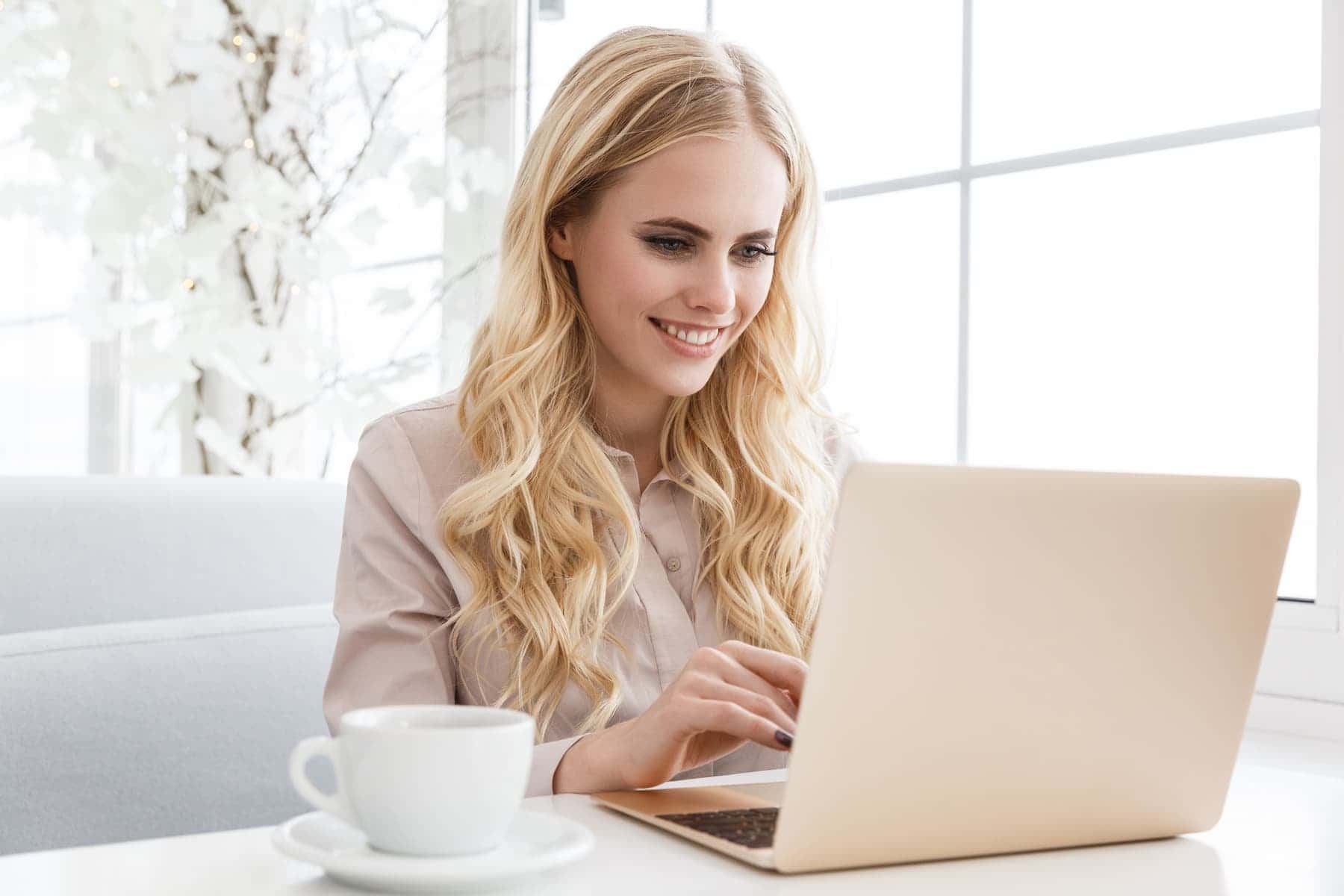 A blonde-haired woman smiles while typing on a laptop, with a cup of coffee beside her.
