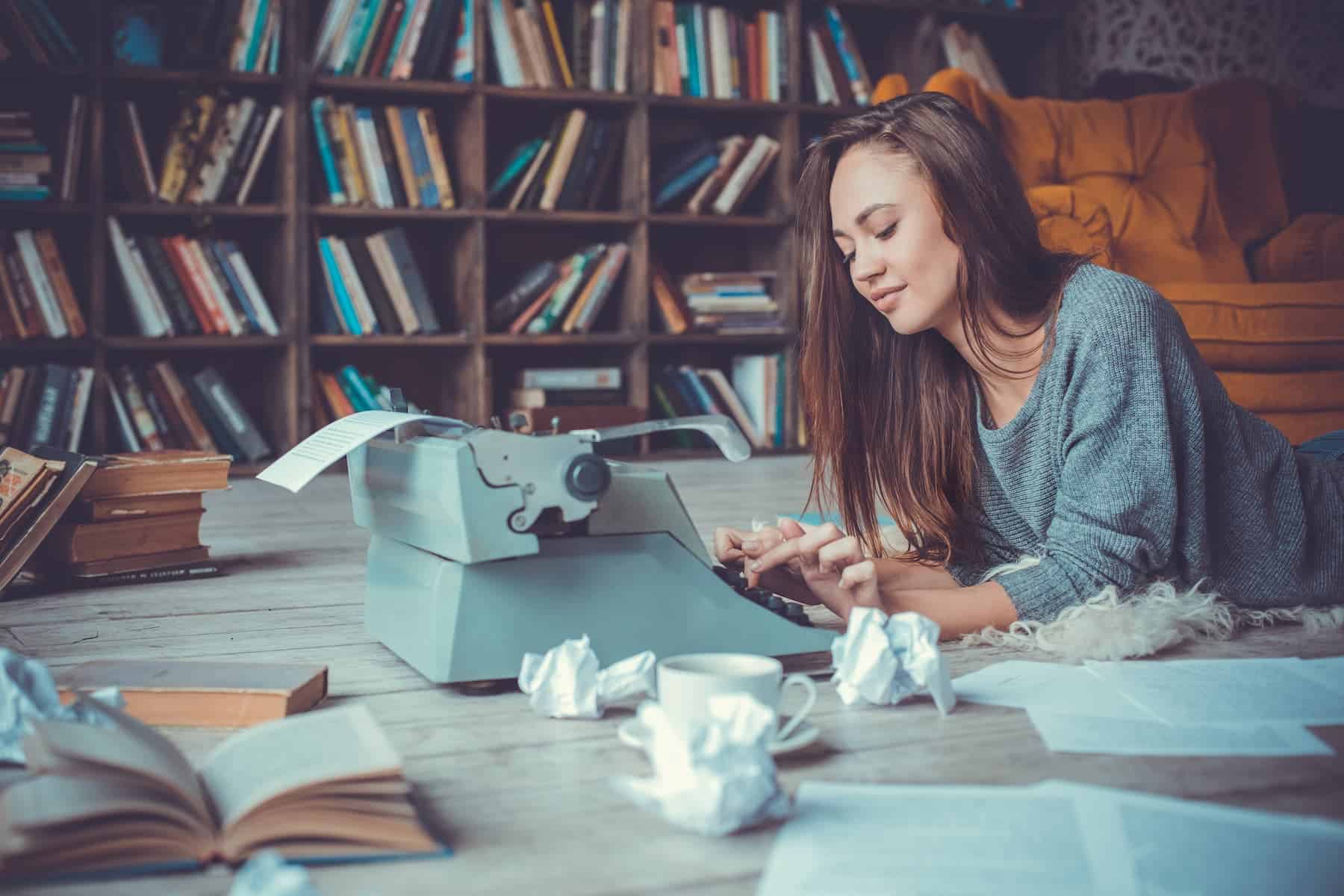 A woman lies on the floor by a vintage typewriter, surrounded by books and crumpled paper, with a bookshelf behind her.