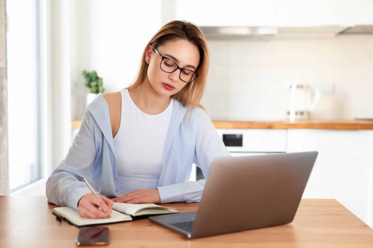 A woman in glasses writes in a notebook at a table with a laptop and phone, with a kitchen counter and kettle in the background.