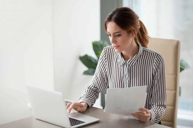 A woman in a striped shirt sits at a desk, reviewing documents while typing on a laptop.
