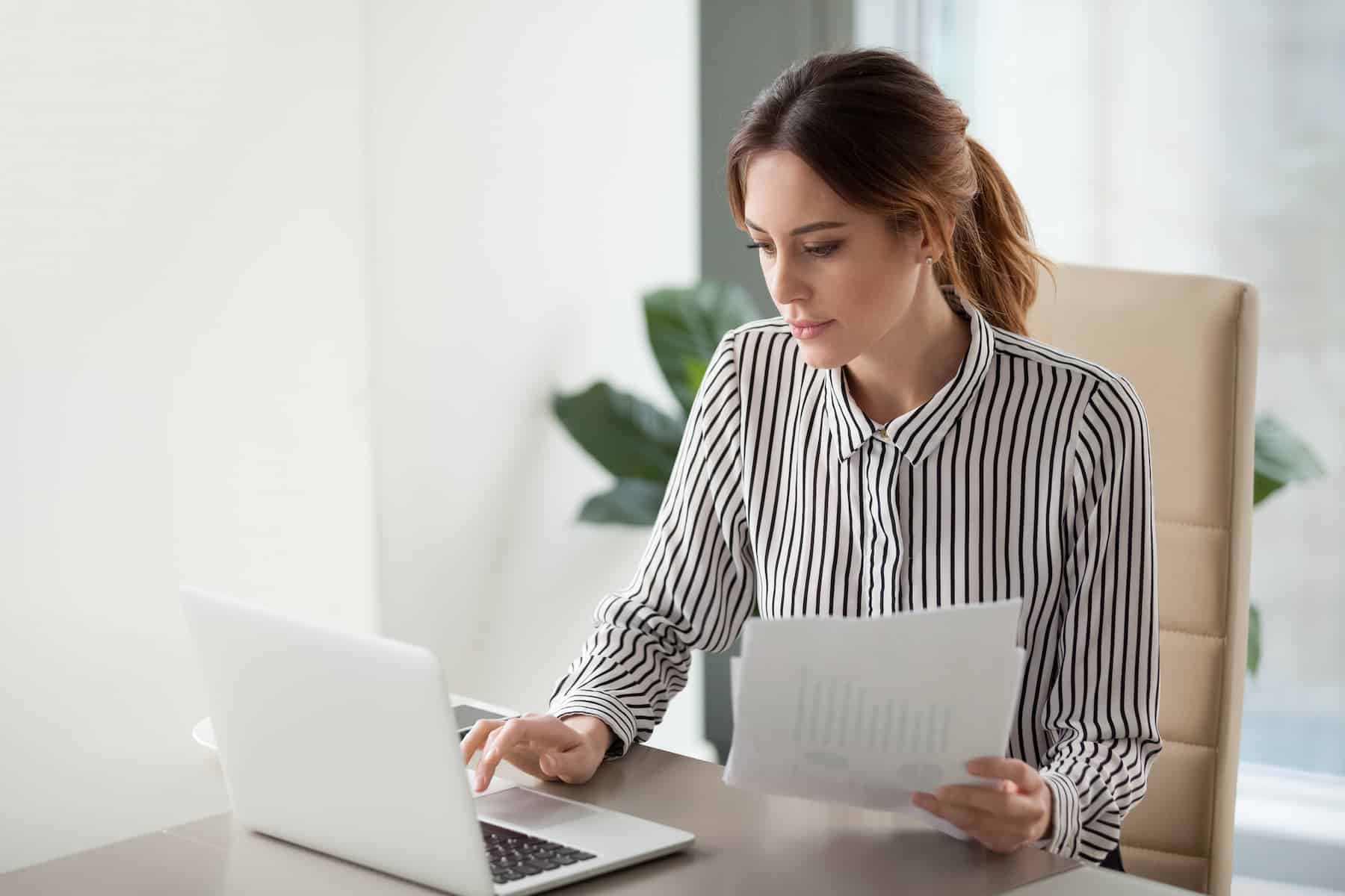 A woman in a striped shirt sits at a desk, reviewing documents while typing on a laptop.