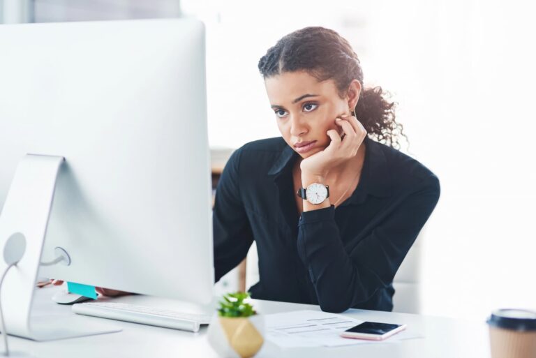A woman at work rests her chin on her hand while looking at a computer screen with a bored expression.