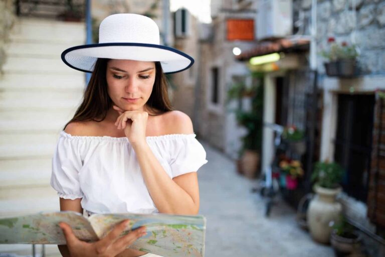 A woman in a white hat and off-shoulder top looks down at a map while traveling, resting her chin on her hand.