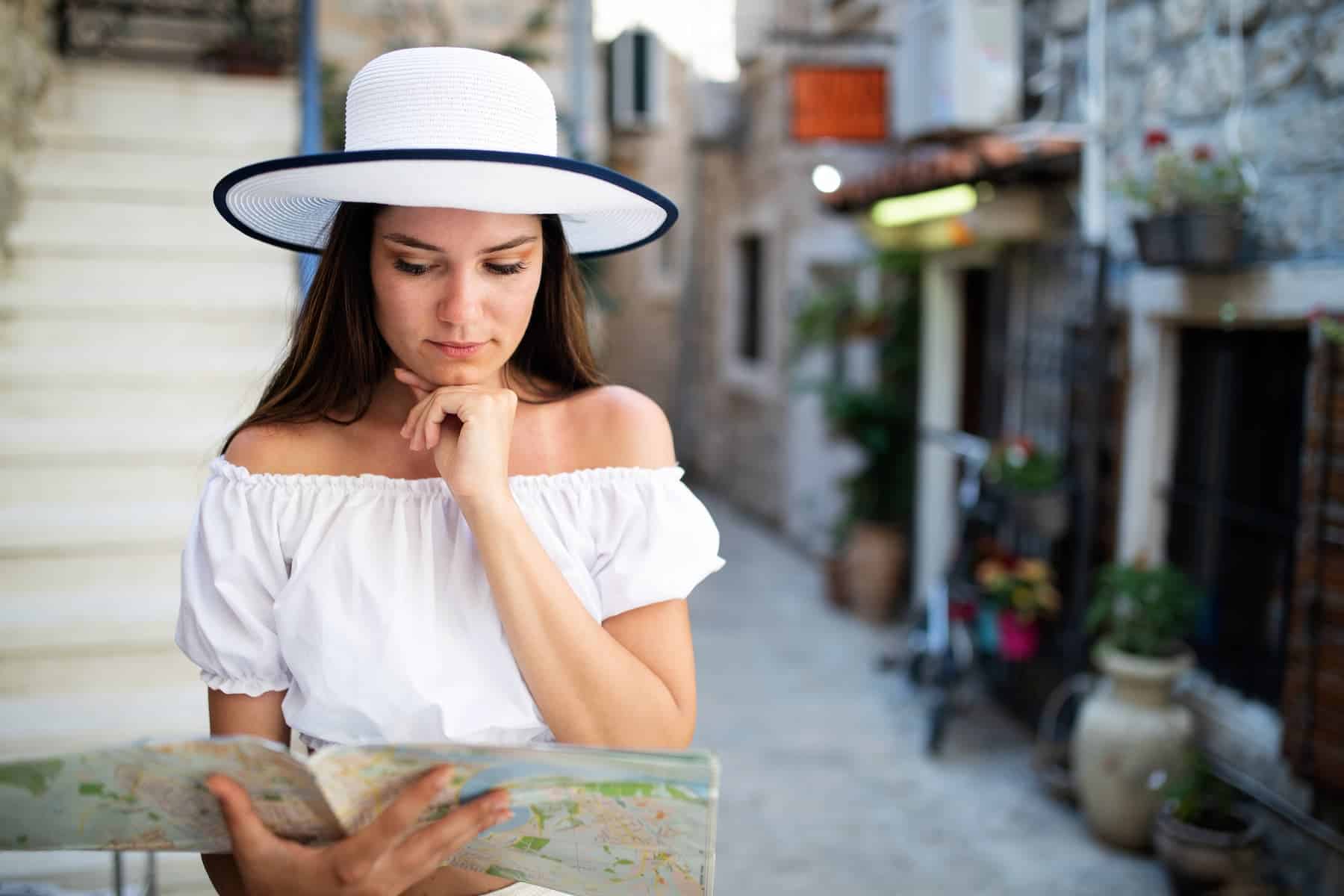 A woman in a white hat and off-shoulder top looks down at a map while traveling, resting her chin on her hand.