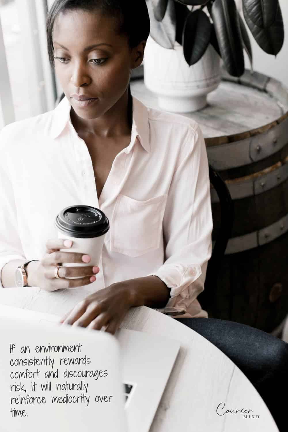 Woman in pink shirt holding coffee while looking down at her laptop in a quiet workspace.