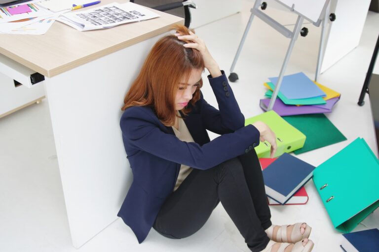 A woman in a blazer sits on the floor with her head in her hands, surrounded by scattered papers, looking defeated.