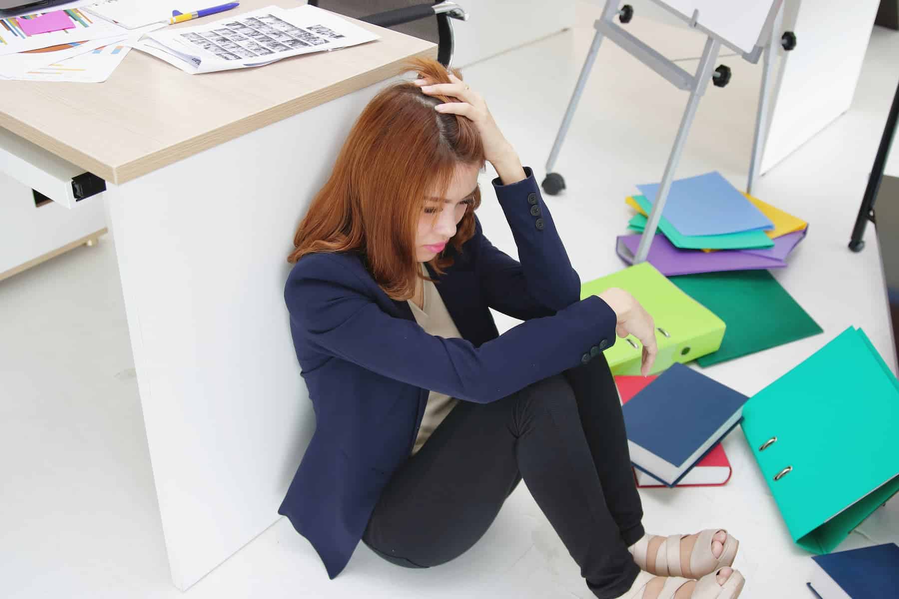 A woman in a blazer sits on the floor with her head in her hands, surrounded by scattered papers, looking defeated.