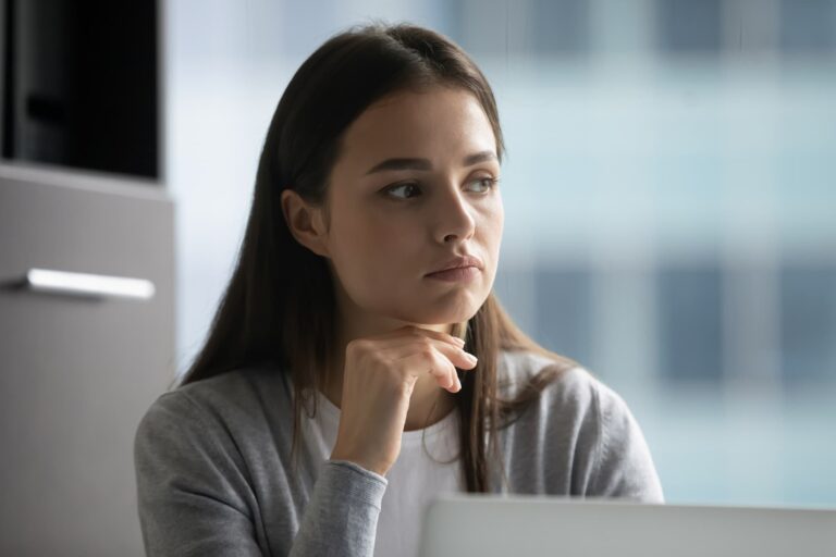A woman wearing a grey cardigan rests her chin on her hand as she looks thoughtfully into the distance.