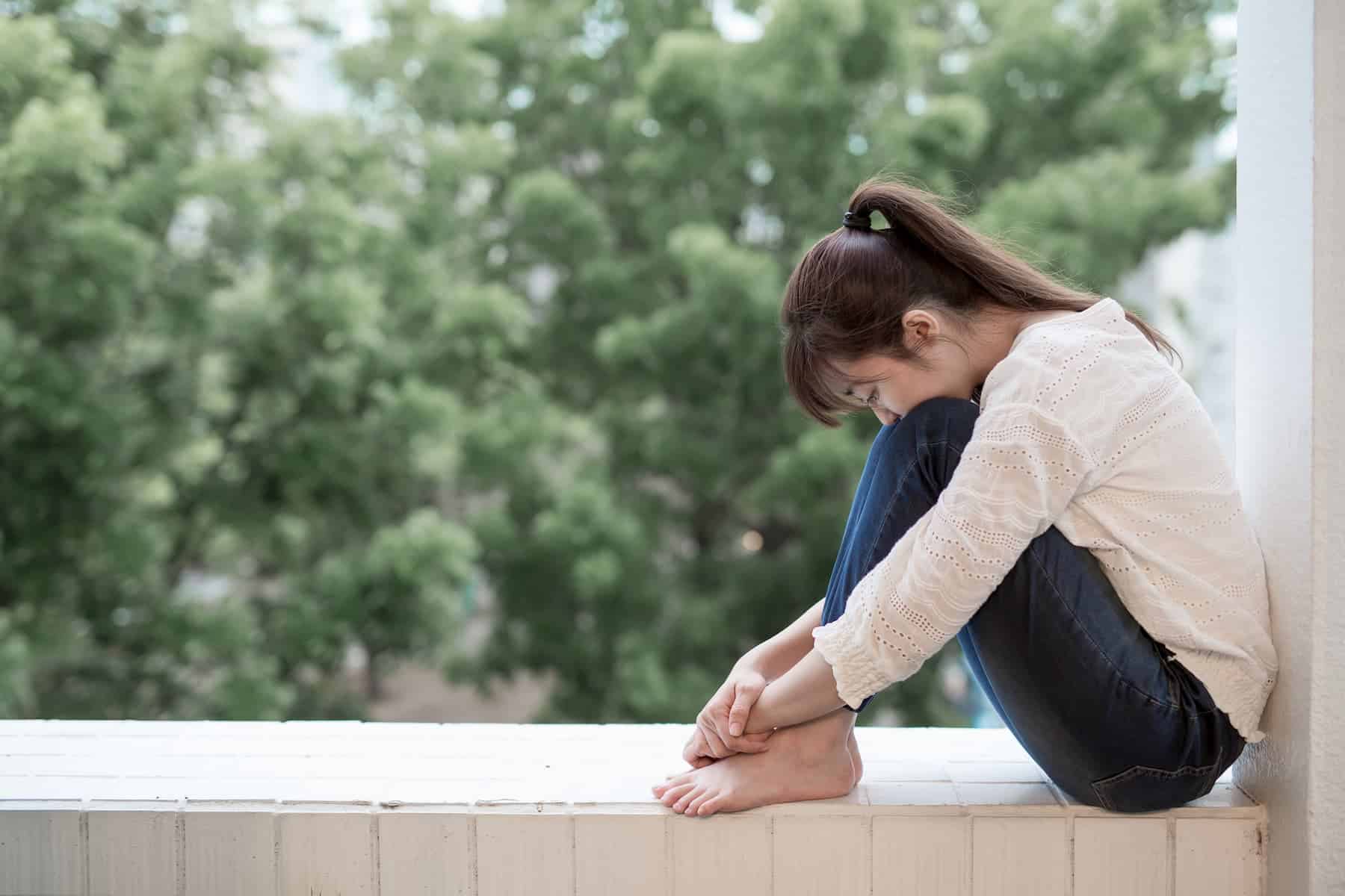 A woman sitting quietly on a ledge with her knees drawn up and her head resting on her knees, with green trees in the background.