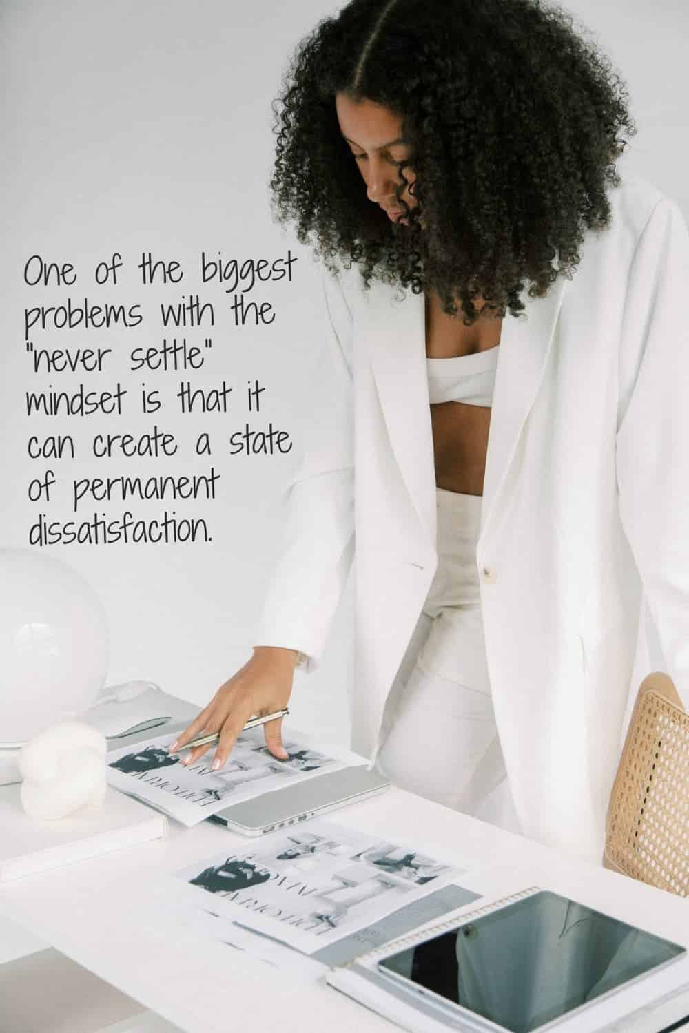 A woman in a white suit jacket looks down at magazines on a white desk while holding a pen.