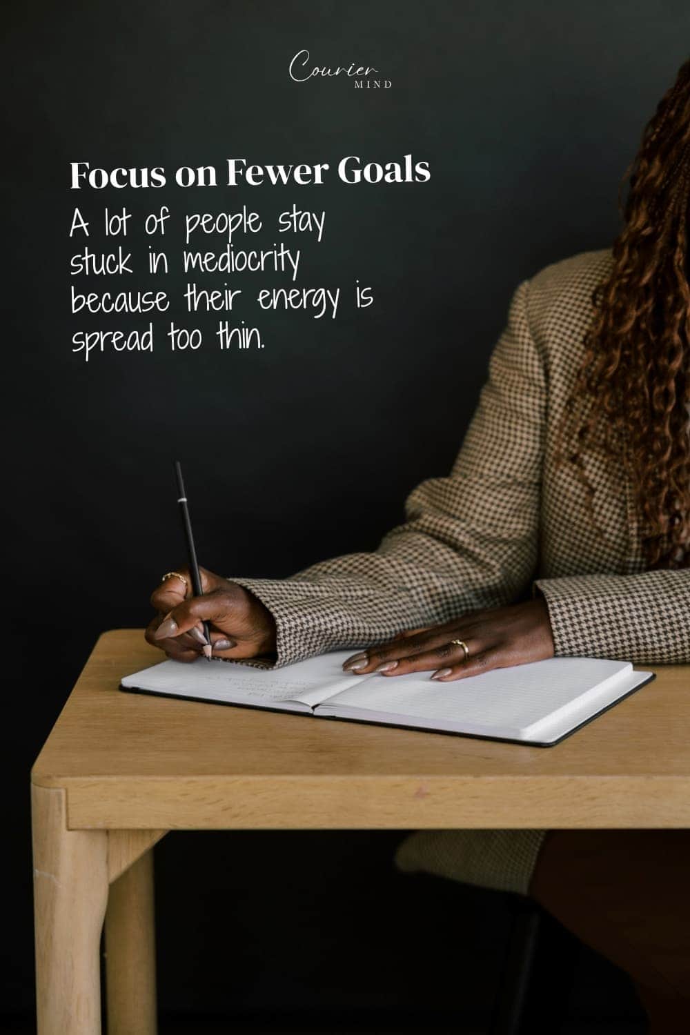 Woman wearing a patterned jacket writing in an open notebook with a pen at a wooden desk against a dark background.