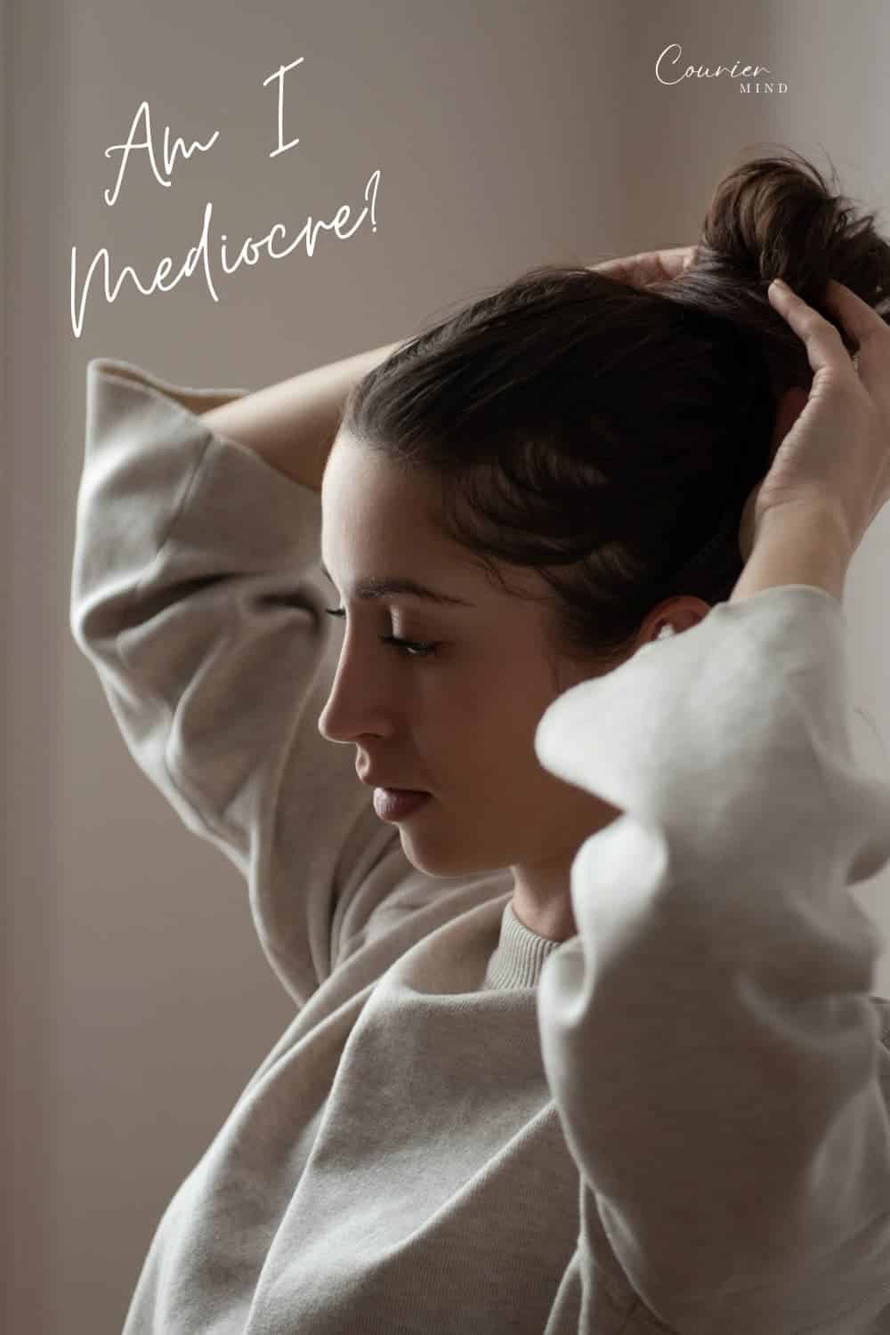 Woman with dark hair pulling her hair into a bun while wearing a light-colored sweatshirt.
