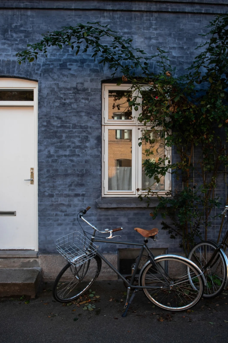 A vintage bicycle with a basket and leather seat is parked against a dark blue brick building.