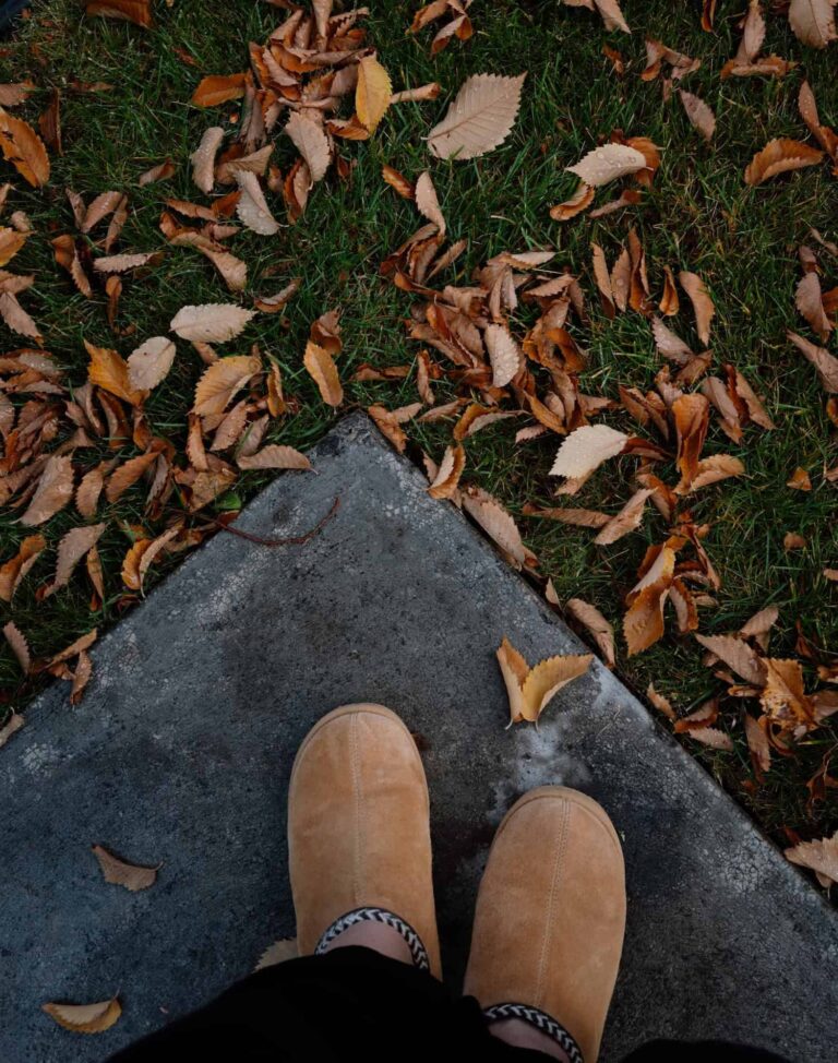 An overhead view captures feet in tan slippers standing on a concrete path with fallen autumn leaves.