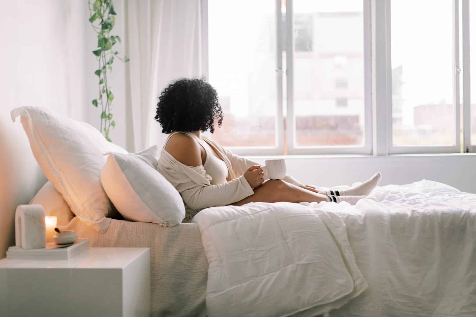 Woman looking out window while sipping coffee in bed.